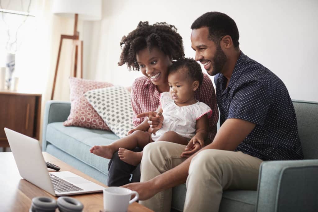 A smiling couple sits on a couch with their young child, looking at a laptop together in a cozy living room. The child sits on one parent's lap while the family enjoys their time together.