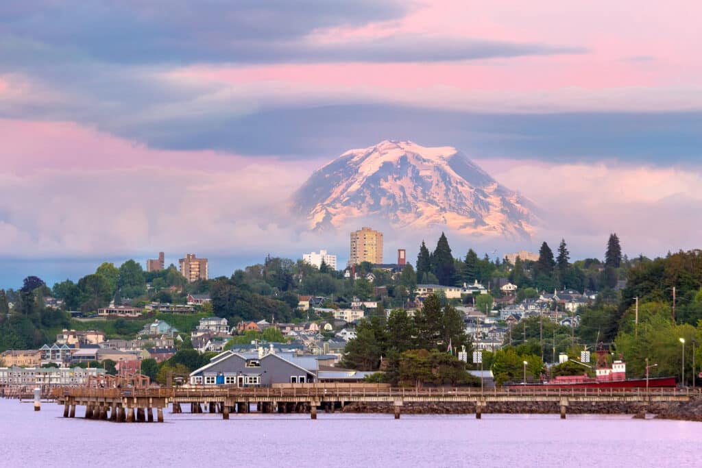 A picturesque waterfront town with a dock and buildings sits beneath a snow-capped mountain, partly shrouded in clouds, under a pastel sky at sunset.