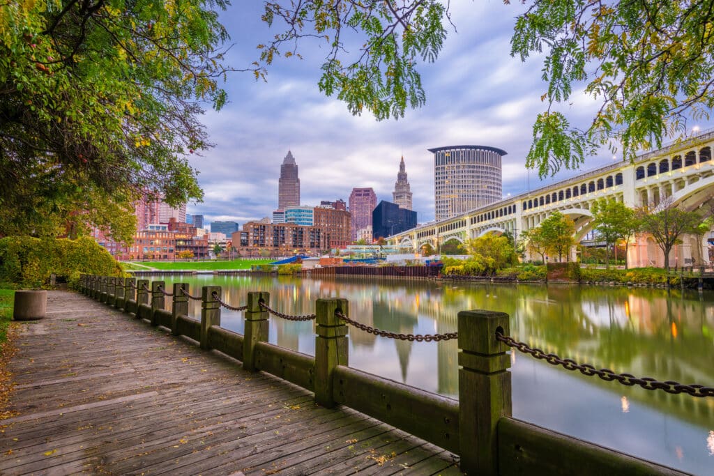 A wooden boardwalk by a calm river with trees on the sides, overlooking the Cleveland city skyline and a large bridge under a partly cloudy sky.