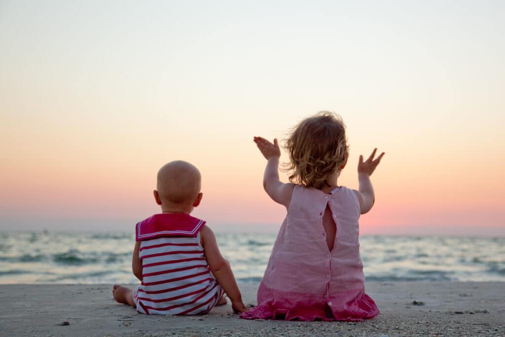 Two young children sit on a sandy beach facing the ocean at sunset. One child is raising their arms while the other sits quietly. Both wear sleeveless outfits and look toward the colorful sky.