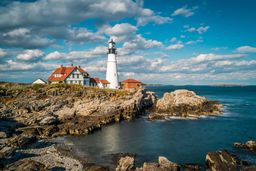 A white lighthouse and red-roofed buildings sit on a rocky coastline under a blue sky with scattered clouds, overlooking the ocean. Waves lap against the rocks in the foreground.