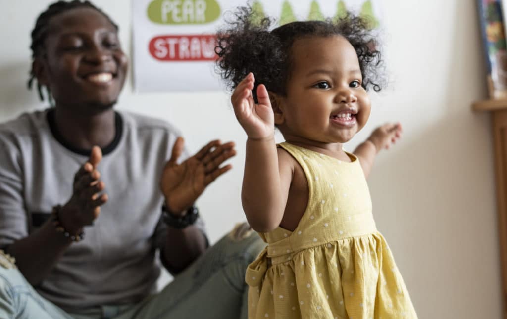 A smiling toddler in a yellow dress raises her arms while a man sitting behind her claps and smiles. A colorful poster is visible on the wall in the background.