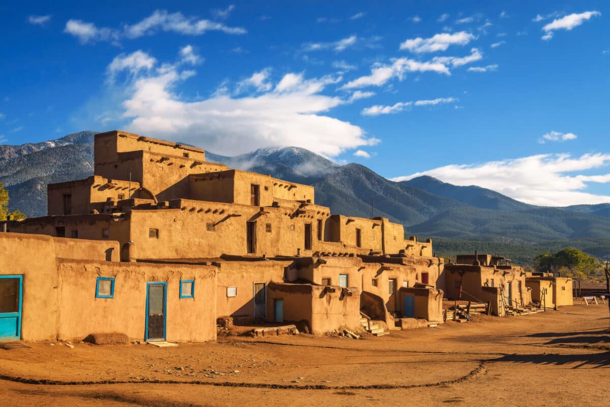 An adobe pueblo with flat roofs and blue doors stands under a bright blue sky, with tree-covered mountains and scattered clouds in the background. The foreground shows a brown dirt ground.