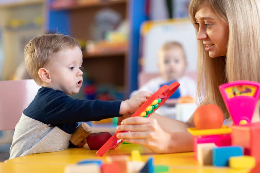 A young child and an adult woman play with colorful educational toys at a table in a bright, cheerful room. The woman smiles as the child reaches toward a toy, encouraging learning and interaction.