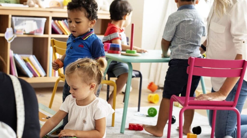 Young children play with toys and blocks in a brightly lit classroom, supervised by an adult. Shelves with books and colorful chairs are visible in the background.