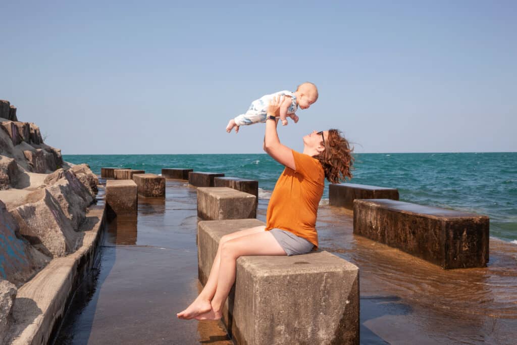 A woman in an orange shirt sits on a concrete block by the sea, holding a baby in the air and smiling. The background shows blue water and sky, with more concrete blocks and rocky formations nearby.