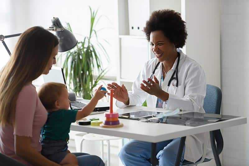 A doctor smiles and interacts with a baby sitting on a woman's lap in a bright office. The baby is playing with a stacking toy while the doctor and mother watch, creating a warm and friendly atmosphere.