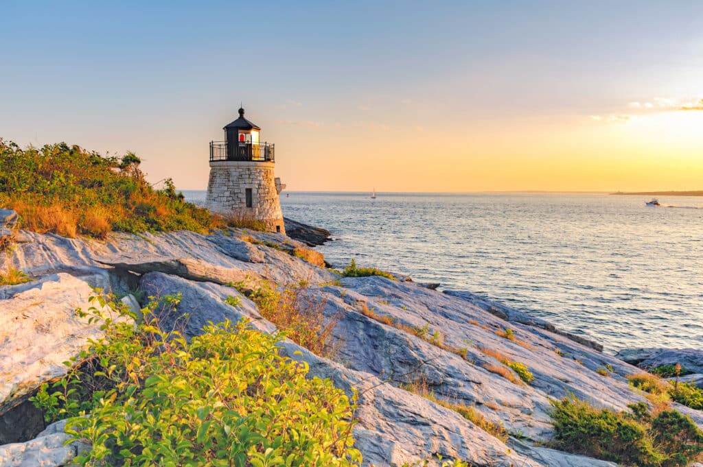 A stone lighthouse stands on rocky cliffs overlooking the ocean at sunset, with warm sunlight illuminating the scene and gentle waves visible in the background.