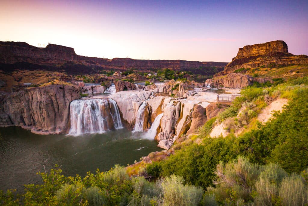 A scenic view of Shoshone Falls cascading over rocky cliffs into a river, surrounded by green shrubbery and plateaus, under a pastel-colored sunset sky.