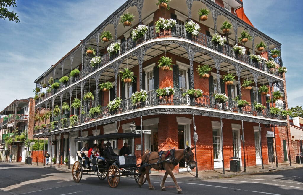 A horse-drawn carriage passes a historic red-brick building with ornate iron balconies and lush hanging plants on a sunny street corner in New Orleans’ French Quarter.