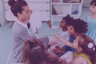 A woman reads a book to a group of young children sitting around her, all appearing engaged and excited in a brightly lit classroom or library setting.