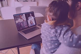 A woman holding a young child sits at a table, participating in a video call on a laptop with four other people visible on the screen.