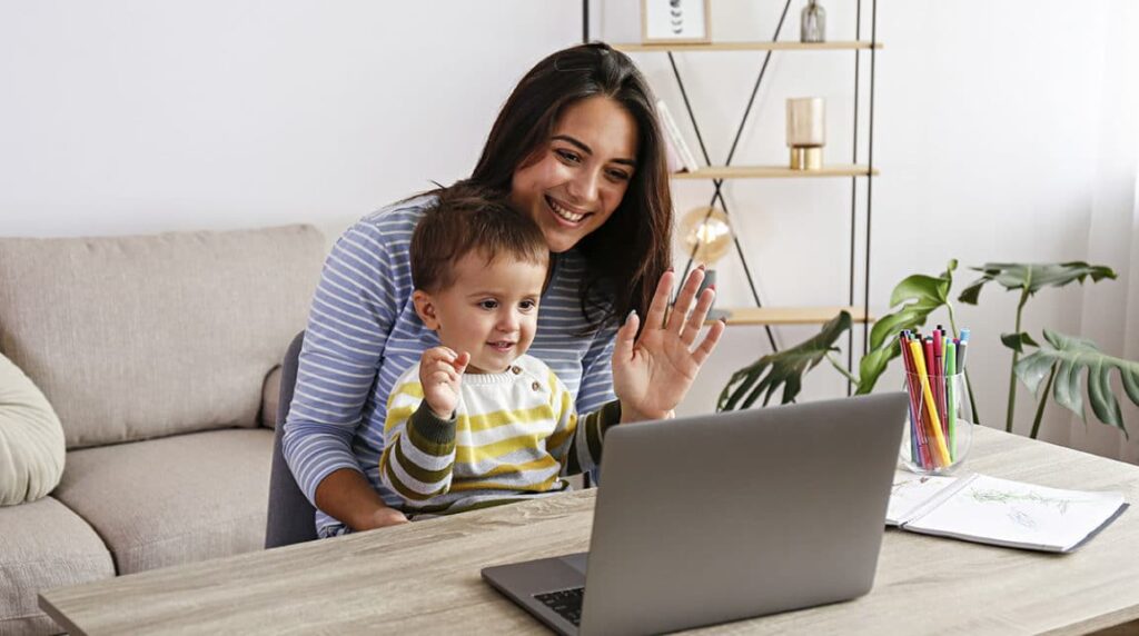 A smiling woman and a toddler sit at a table, waving at a laptop screen during a video call. The room has a sofa, plants, and shelves in the background, with colored pencils and paper on the table.