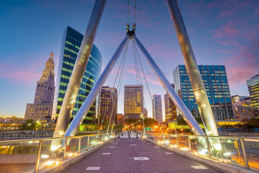 Pedestrian bridge with modern cables leads toward a city skyline at sunset, featuring tall glass buildings and a historic clock tower under a colorful sky in Hartford, Connecticut.