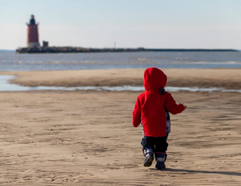 A small child in a red jacket walks alone on a sandy beach toward the water, with a lighthouse visible in the distance on a jetty.