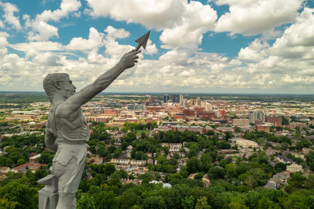 A large statue of a man holding a spear overlooks a cityscape with tall buildings, green trees, and residential areas under a sky filled with white clouds.