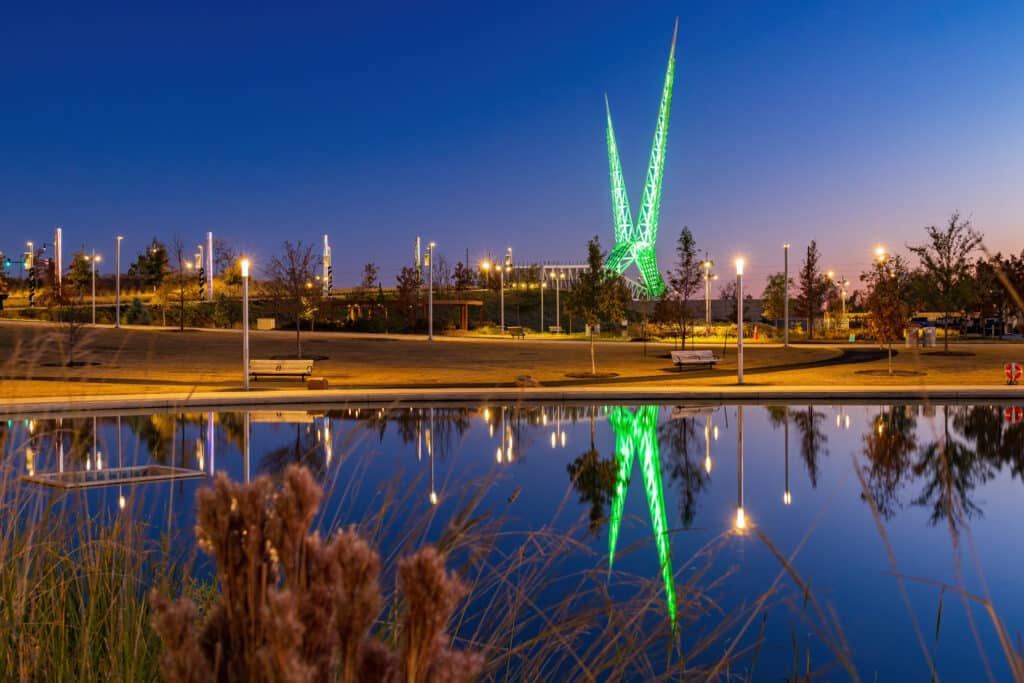 A nighttime scene in a park features a reflective pond, illuminated benches, and a striking sculpture with two green-lit spikes. City lights and trees are visible under a deep blue sky.
