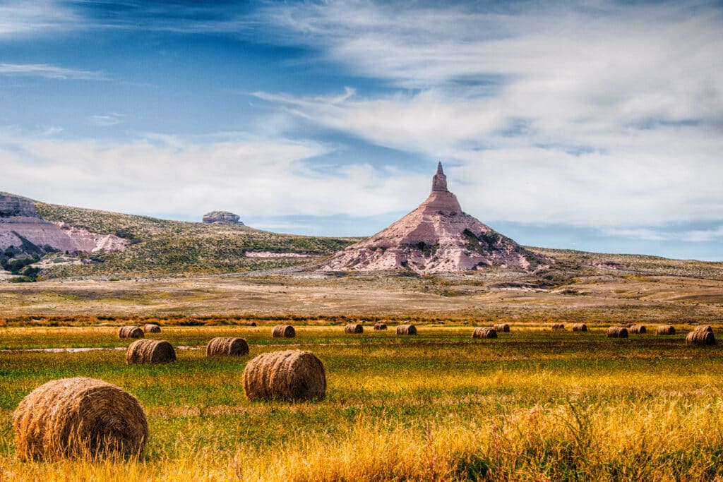 Round hay bales scattered across a grassy field, with a prominent, rocky butte rising in the background under a partly cloudy blue sky.