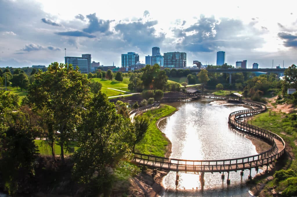 Winding boardwalk over a river in a lush park, with a city skyline and tall buildings in the background under a partly cloudy sky.