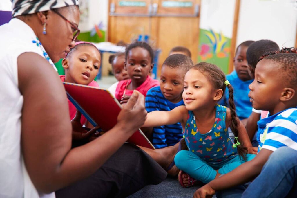 A woman reads a book to a group of young children sitting on the floor. The children are engaged and looking at the book, with one girl reaching out toward it. The setting appears to be a classroom or daycare.