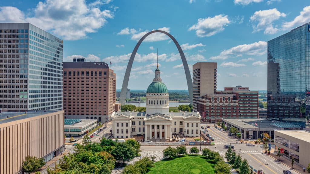 A cityscape of St. Louis featuring the historic Old Courthouse in the foreground, the Gateway Arch in the background, and modern office buildings on either side under a blue sky with scattered clouds.