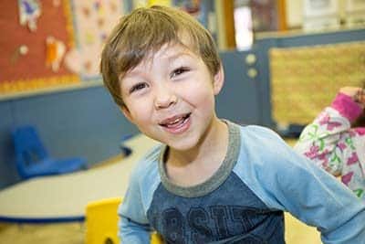 A young child with light brown hair smiles brightly at the camera in a colorful classroom setting.
