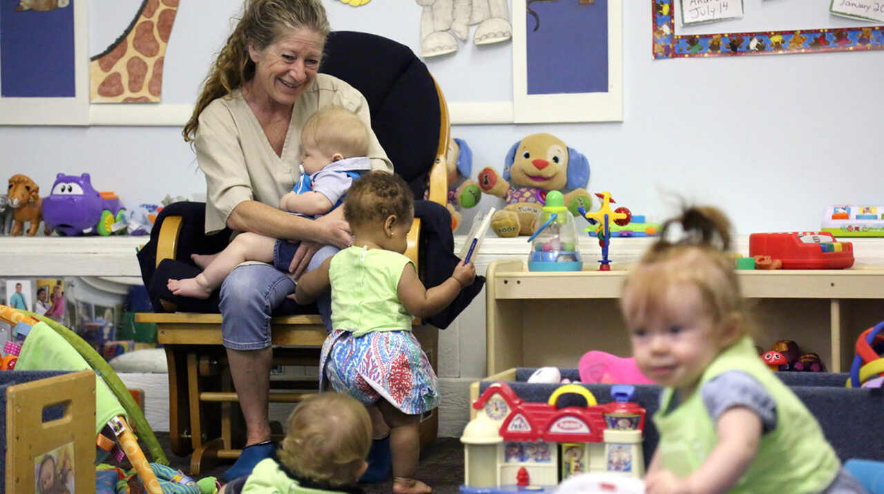 A woman sits in a chair holding a baby in her lap, smiling, while three other young children play on the floor with toys in a colorful daycare room.