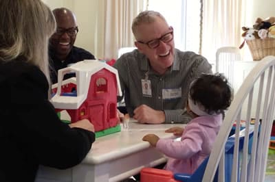 Two adults and a toddler sit around a small table playing with a toy barn. Everyone appears to be smiling and engaged, creating a warm, friendly atmosphere in a brightly lit room.