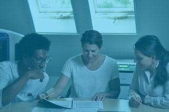 Three people sit around a table, reviewing documents together and holding pencils. They appear to be working or studying collaboratively in a well-lit room with two windows.
