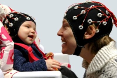 A baby and an adult, both wearing black caps with sensors and wires for brain activity monitoring, face each other and smile warmly. The baby sits in a high chair, and the adult leans in close.
