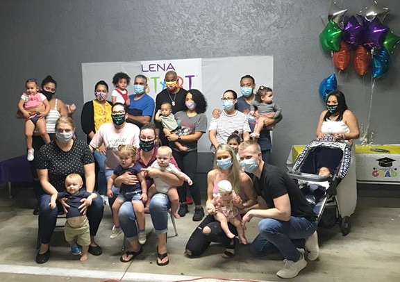 A group of adults and young children, most wearing face masks, pose together indoors in front of a sign and colorful balloons. Some adults are holding babies, and there is a stroller with a baby on the right.