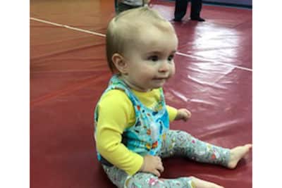 A young child with blond hair, wearing a yellow shirt and colorful overalls, sits on a red gym mat, looking forward and smiling slightly.