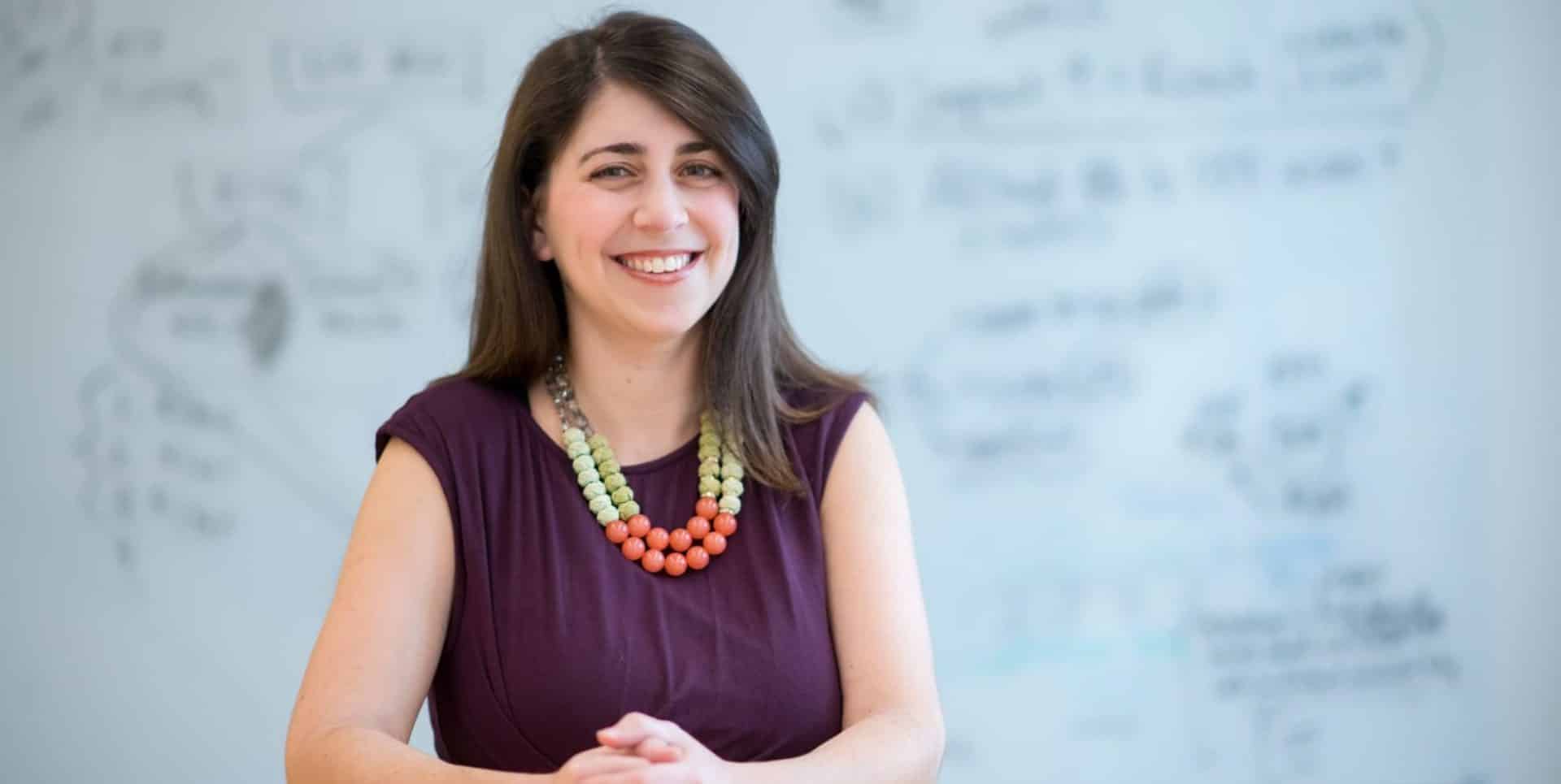 A woman with long brown hair, wearing a maroon dress and a colorful beaded necklace, smiles while sitting with her hands clasped. A whiteboard with writing and diagrams is blurred in the background.