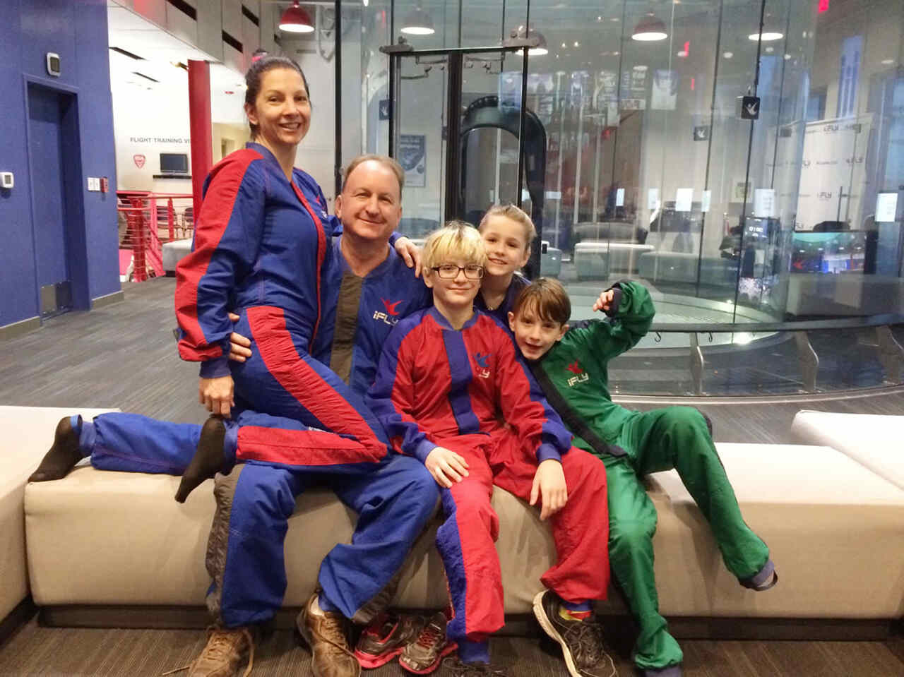 A smiling family of five in colorful flight suits sits on a bench inside an indoor skydiving facility, with the wind tunnel visible in the background.