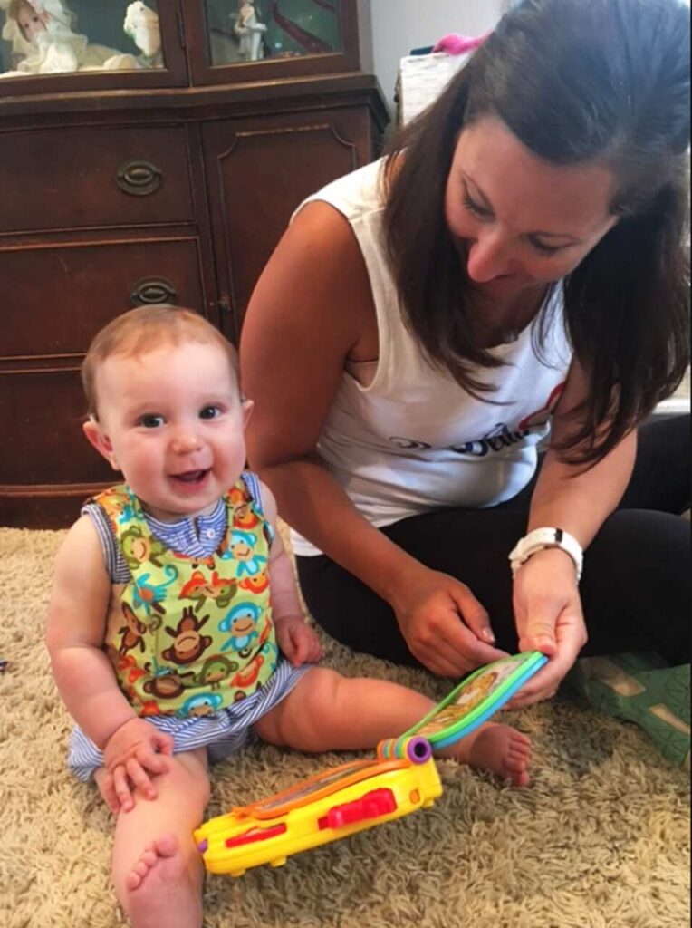 A smiling baby sits on a carpet next to a woman who is reading a children's book. The baby wears a colorful bib with animal designs, and a toy is on the floor. The woman looks at the book and smiles warmly.