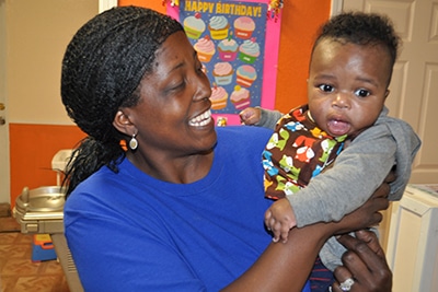 A woman in a blue shirt smiles while holding a baby wearing a colorful bib. The baby looks surprised. In the background, there is a “Happy Birthday” sign with cupcakes.