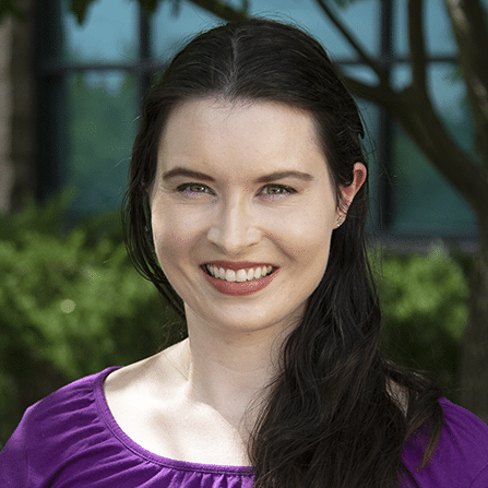 A woman with long dark hair wearing a purple top smiles at the camera outdoors, with greenery and a building window in the background.