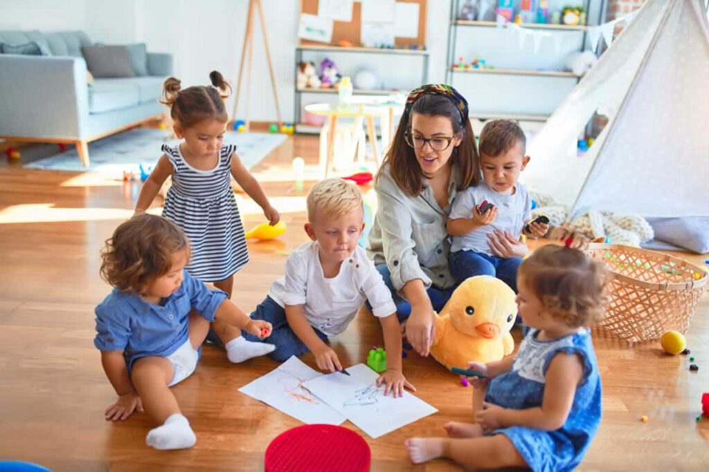 A woman sits on the floor with five young children, engaging in drawing and playing with toys in a bright, cozy room with a teepee and scattered play items.