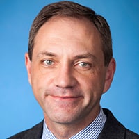 A man with short brown hair wearing a dark suit jacket, light blue dress shirt, and striped tie, smiling slightly against a plain blue background.