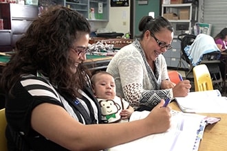 Two women sit at a table smiling and writing in notebooks; one holds a baby on her lap. The room has shelves, books, and classroom materials in the background.