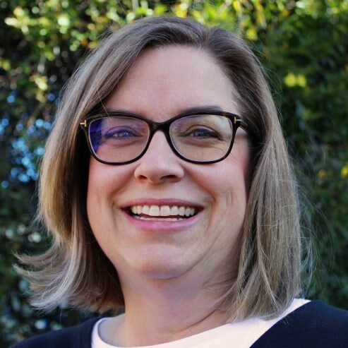 A woman with shoulder-length light brown hair and glasses smiles outdoors. She is wearing a white top and a dark sweater, with greenery blurred in the background.