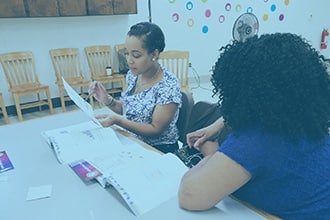 Two women sit at a table looking over papers and documents. Chairs are lined up against the wall in the background, and colorful circles decorate the wall. One woman is holding a sheet of paper and a pen.