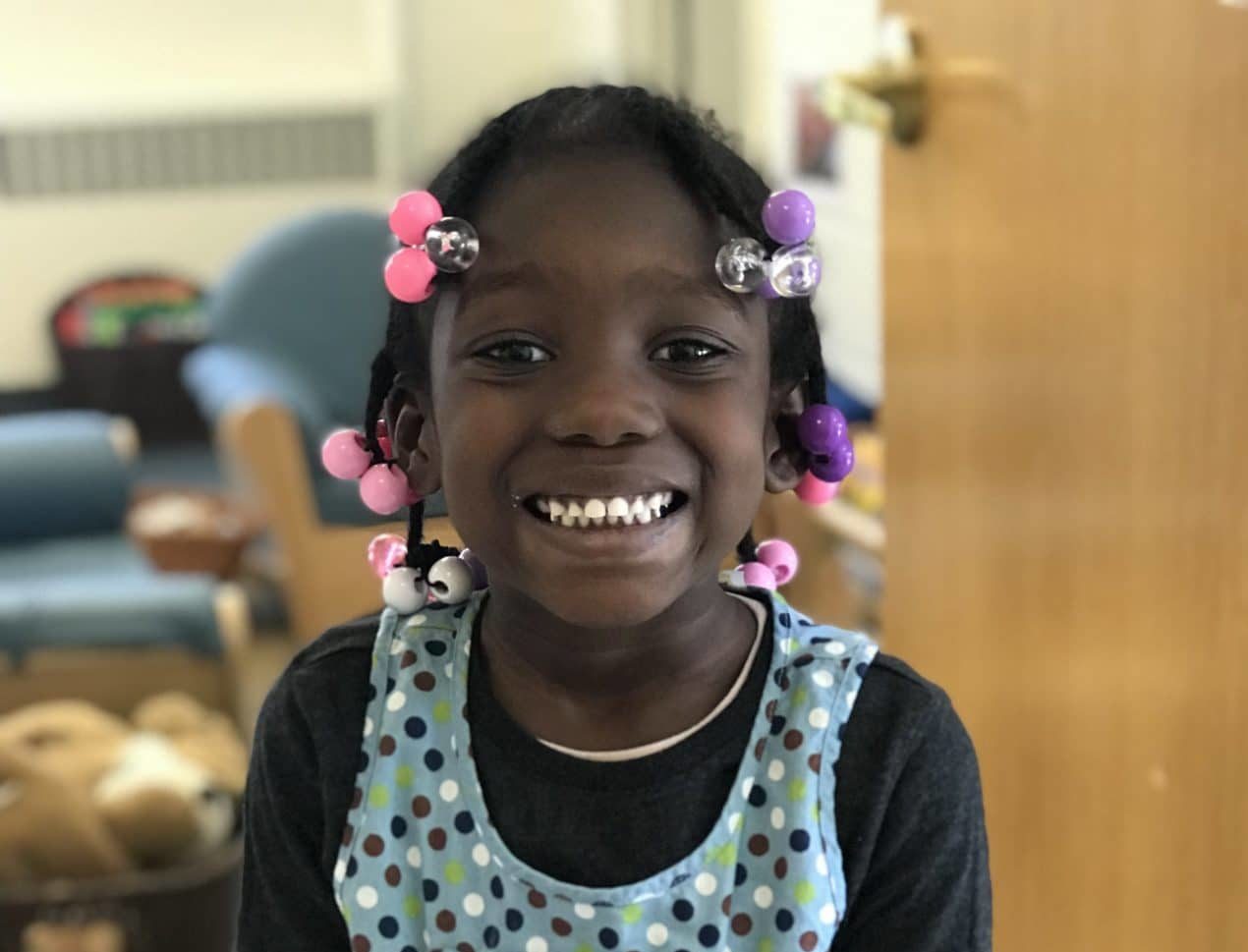 A young girl with braided hair decorated with colorful beads smiles brightly. She is wearing a polka-dot apron over a dark shirt and is standing in a room with toys and chairs in the background.