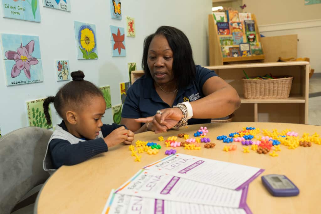 A woman helps a young girl with colorful counting manipulatives at a round table in a classroom decorated with flower artwork and bookshelves. Educational papers and a timer are also on the table.