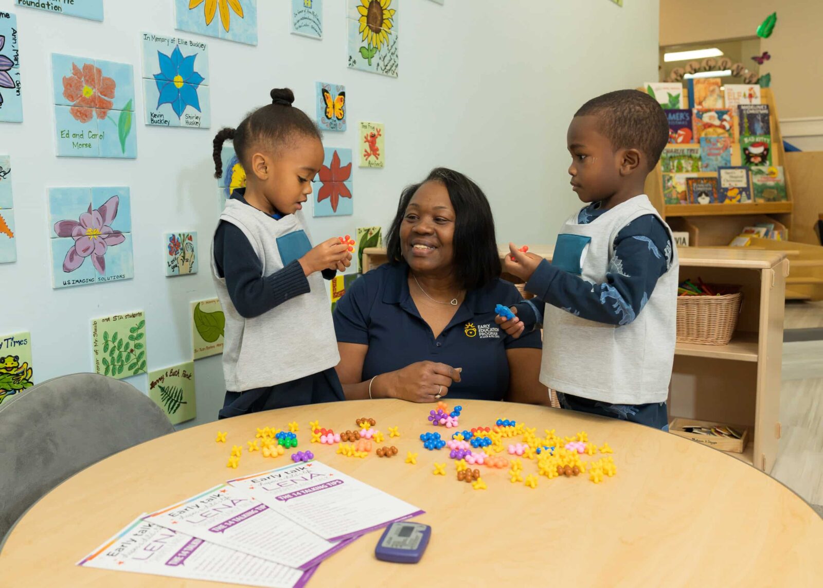 A teacher sits at a round table with two young children, who are playing with colorful star-shaped counters. Educational materials and flower artwork decorate the classroom walls in the background.