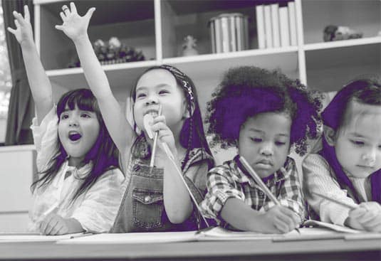 Four children sit at a table in a classroom. Two are smiling and raising their hands enthusiastically, while the other two are focused on writing or drawing. Shelves with books and toys are in the background.