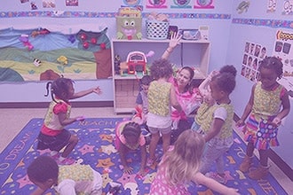 A group of young children and a teacher play and interact energetically in a colorful classroom decorated with artwork, toys, and educational materials on the walls and shelves.