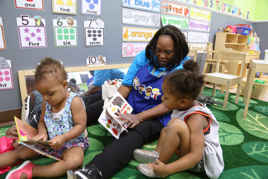 A teacher sits on a green carpet with two young girls in a colorful classroom, reading books together. Educational posters and toys decorate the walls and shelves in the background.