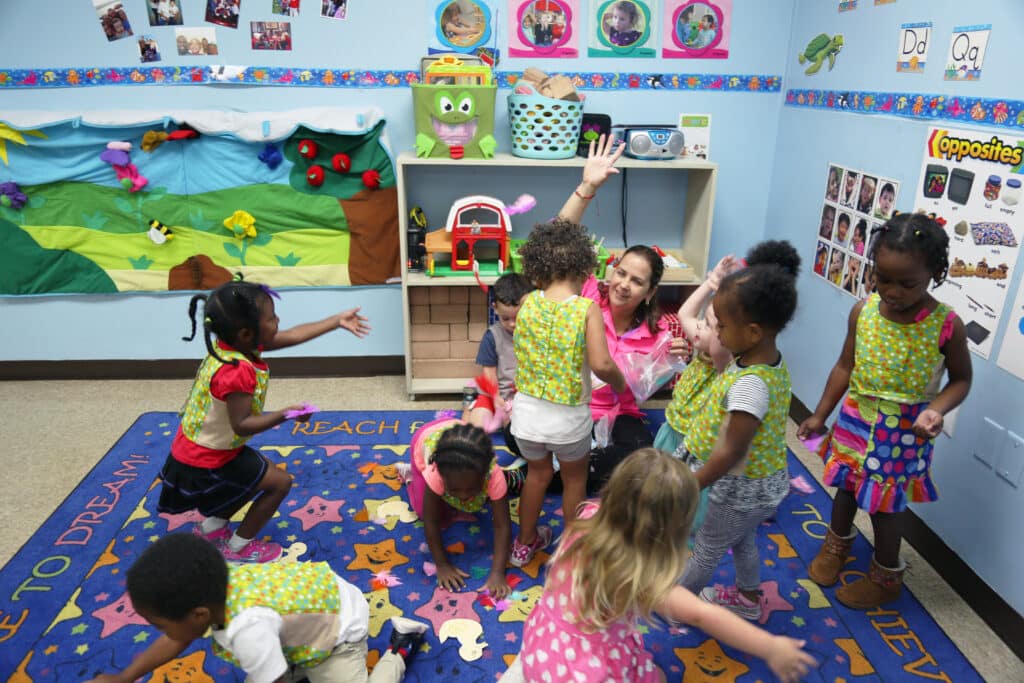 A group of young children and a teacher play and interact energetically in a colorful classroom decorated with educational posters and crafts. The teacher smiles, raising her hand, while kids move around on a vibrant alphabet rug.