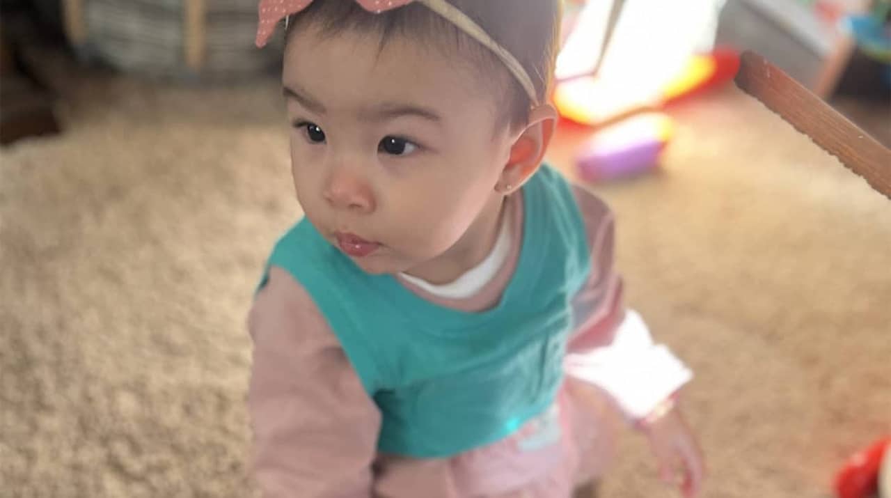 A young toddler wearing a pink headband with a bow and a teal bib looks to the side while standing on a carpeted floor in a softly lit room.